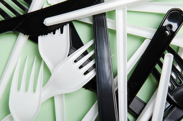 Plastic white and black forks and knives on a green background. Concept plastic dishes, plastic pollution. Flat lay, top view, texture - Photo, Image