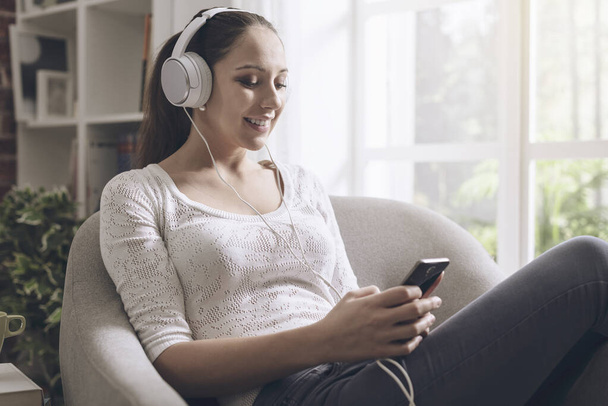Happy young woman listening to music at home, she is connecting with her smartphone and wearing headphones - Fotó, kép