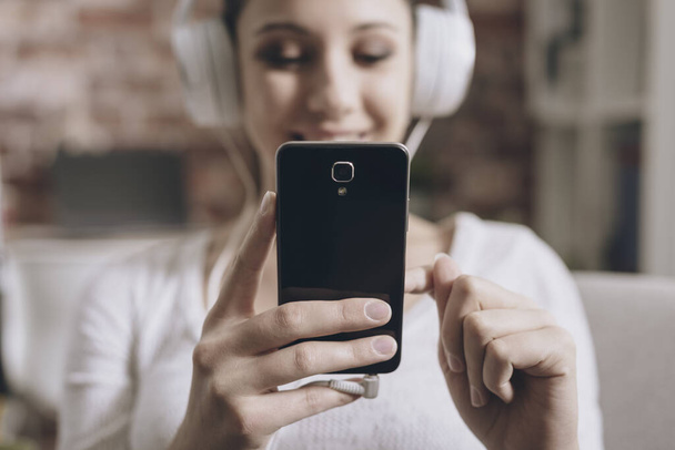 Ευτυχισμένος νεαρή γυναίκα να ακούτε μουσική στο σπίτι, είναι σύνδεση με smartphone της και φορώντας ακουστικά - Φωτογραφία, εικόνα