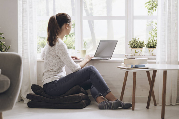 Young cute woman connecting with her laptop at home, she is sitting on the floor and typing - Foto, Imagem
