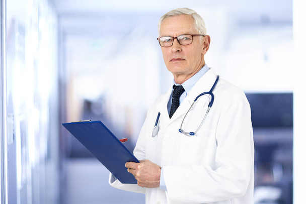 Portrait shot of elderly male doctor holding clipboard in his hand while standing in the hospital. - Foto, imagen