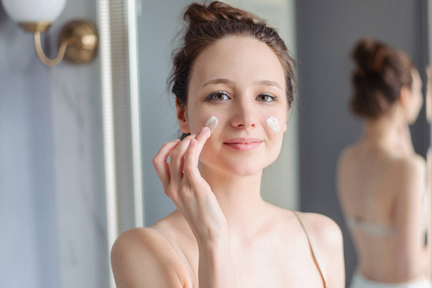 Young girl applies cream to face. Home light bathroom, lifestyle, natural skin care, love yourself. Cream on cheeks. A woman without makeup, natural. - Foto, Imagem