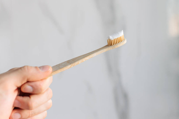 Eco-friendly wood and bamboo toothbrush with paste applied on a bright white marble background in your hand. Care of teeth. - Fotografie, Obrázek