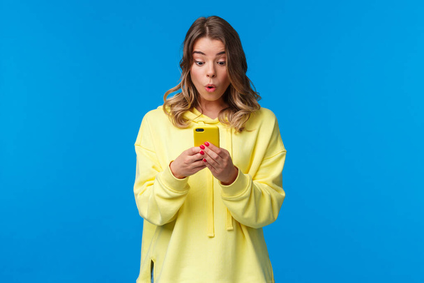 Удивлена и заинтригована симпатичная блондинка, видящая что-то интересное в Интернете, просматривающая поисковую одежду в магазине одежды, использующая крутое новое приложение, держащая смартфон
 - Фото, изображение