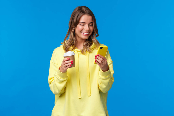 Zorgeloos vrolijke Europese vrouw in gele hoodie, het houden van take-away koffie en surfen op internet in mobiele telefoon, sms 'en vriend lachen over grappige video of meme, blauwe achtergrond - Foto, afbeelding