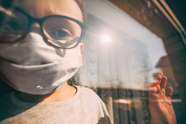 a child in quarantine because of a virus sits at home in a mask and looks out the window on a sunny day - Fotografie, Obrázek