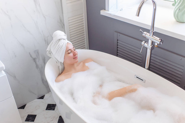 A young girl with a turban - a towel - on her head lies in a beautiful bright home bathroom and relapses. Daylight, marble tiles. The woman 's eyes are closed. Home spa and body care, lots of foam. - Photo, image