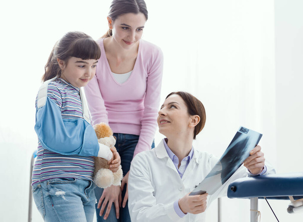 Friendly doctor showing an x-ray image of a broken bone to a young patient, the girl is wearing an arm brace - Foto, Imagen