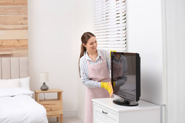 Young chambermaid wiping dust from TV with rag in hotel room - Foto, immagini