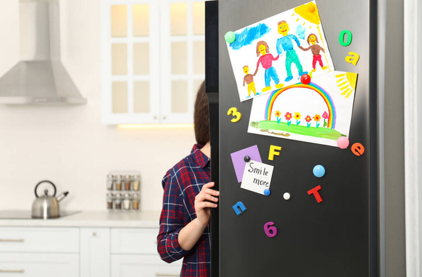 Woman opening refrigerator door with child's drawings, notes and magnets in kitchen. Space for text - Foto, Imagen