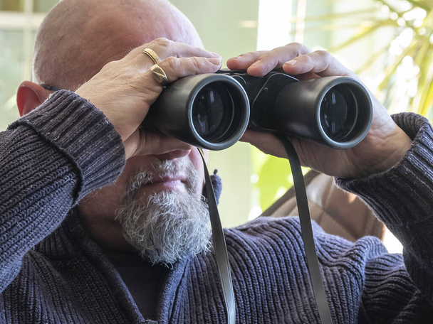 Starší muž sedí, dívá se dalekohledem do skleněných dveří a v dálce něco pozoruje. - Fotografie, Obrázek