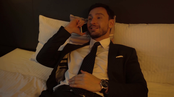 明るいビジネスマンがベッドに横になってホテルのスマートフォンで話してる - 映像、動画