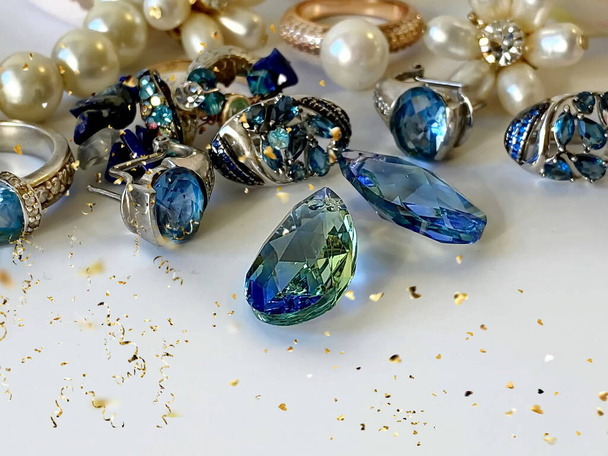 синій кристал і біла перлина Ювелірні вироби золоті срібні сині кам'яні сережки зелений смарагд, сині кришталеві дорогоцінні камені, кільця, намисто крупним планом дробовик, на білому тлі розмиті легкі золоті елементи
  - Фото, зображення