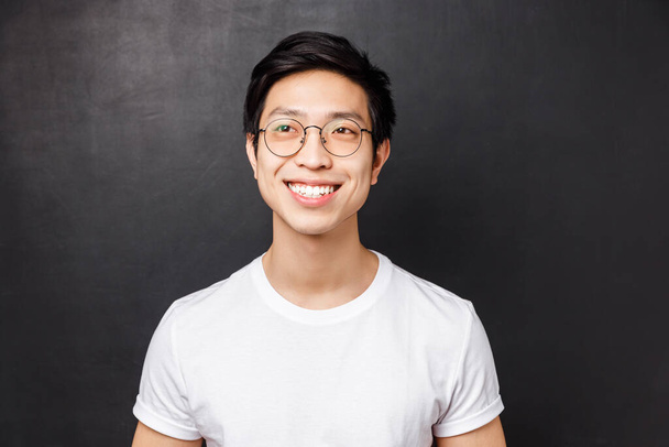 primer plano retrato de soñador feliz joven asiático hombre en blanco camiseta, gafas, radiante sonrisa emocionado, mirando a la izquierda alegre, de pie negro fondo, recordar algo lindo
 - Foto, Imagen