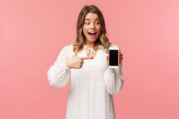 Retrato de mujer rubia emocionada y sorprendida, feliz en vestido blanco, apuntando con el dedo a la pantalla del teléfono móvil, mirando el teléfono inteligente con sonrisa complacida y sorprendida, de pie fondo rosa
 - Foto, imagen