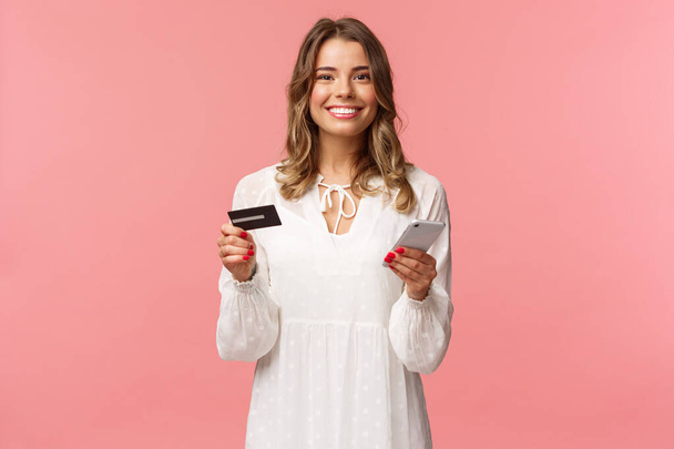 Retrato de mujer rubia sonriente feliz pedir comida en línea, uso de la aplicación para la tienda de Internet de compras, la celebración de la tarjeta de crédito y el teléfono inteligente, sonriendo encantado con la cámara, fondo rosa
 - Foto, imagen