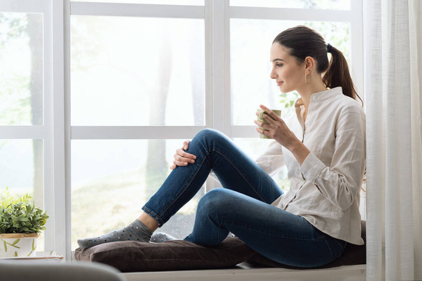 Молода жінка розслабляється вдома біля вікна і має чашку кави
 - Фото, зображення