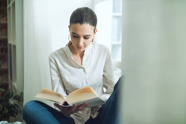 Jonge serene vrouw ontspannen thuis en het lezen van een boek naast een raam - Foto, afbeelding