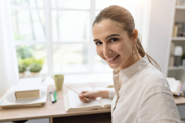 Boldog fiatal nő tanul otthon, ül az asztalnál, és mosolyog a kamerába - Fotó, kép
