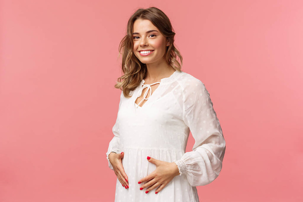 Maternity, women and beauty concept. Tender, cute smiling blond woman in white dress feeling happiness and love, touching belly as expecting child, being pregnant, pink background - Φωτογραφία, εικόνα