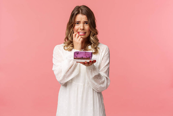 Holidays, spring and party concept. Alarmed, worried young blond girl hesitating eat cake or not, being on diet, trying stay healthy, holding dessert, biting finger nervously, pink background - Photo, Image
