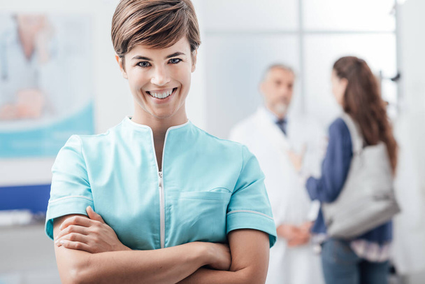 Sonriente enfermera joven posando en la clínica con los brazos cruzados, un médico está dando la bienvenida a un paciente en el fondo
 - Foto, imagen