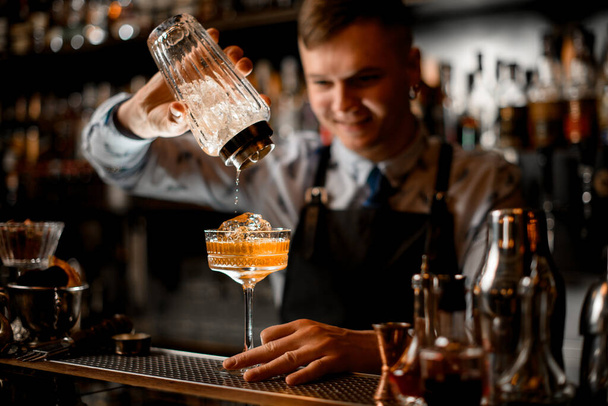 smiling young bartender pours finished cocktail from glassy shaker into glass with ice. - Foto, imagen