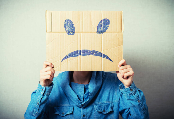 Curly man with a kraft cardboard instead of a head, a sad smiley. On a gray background. - Φωτογραφία, εικόνα