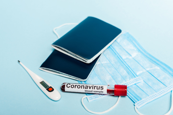tubo de ensayo con muestras de sangre y letras de coronavirus cerca de máscaras médicas, termómetro y pasaportes sobre fondo azul
 - Foto, Imagen
