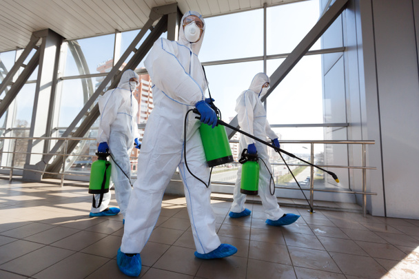 Muži v respirátorech a ochranných oblecích čistí veřejné prostory chemikáliemi - Fotografie, Obrázek