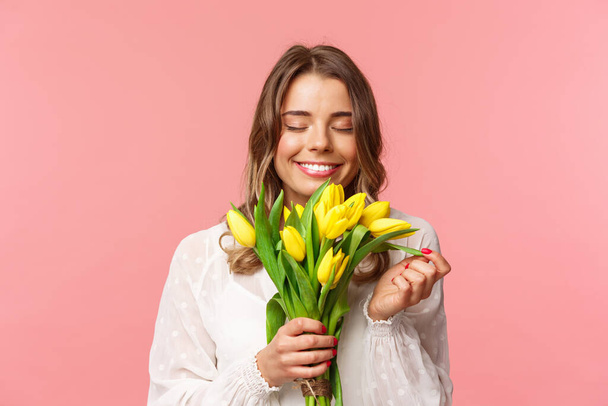 Jaro, koncepce štěstí a oslav. Detailní portrét krásné, romantické blond dívky čichající vůni krásné žluté tulipány, zavřené oči a šťastný úsměv, stojící růžové pozadí - Fotografie, Obrázek