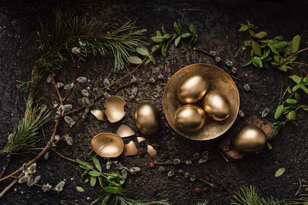 Golden easter eggs are scattered on a dark background with dry green branches. eggs in a golden bowl. - Foto, Bild