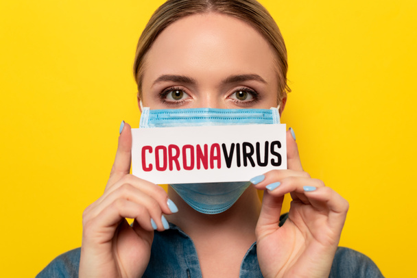 junge Frau in medizinischer Maske mit einem Ausweis mit Coronavirus-Schriftzug auf gelb isoliert - Foto, Bild