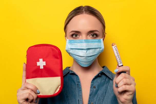 young woman in medical mask holding test tube with coronavirus blood sample and first aid kit on yellow background - Photo, Image