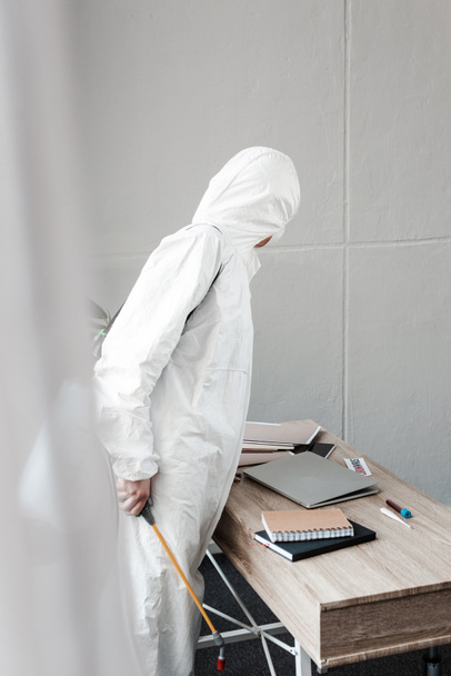 Person in weißem Warnanzug, Atemschutzmaske und Schutzbrille zur Desinfektion des Arbeitsplatzes im Büro, Coronavirus-Konzept - Foto, Bild