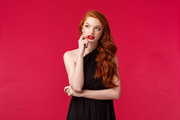 Portrait of thoughtful creative young redhead elegant woman with ginger hair in black dress, biting lip and looking up dreamy, thinking about solution, standing over red background - Фото, изображение