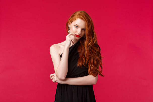Portrait of sassy and seductive young elegant woman with red ginger hair, wearing black dress, evening makeup and red lipstick, look from under forehead daring and flirty, red background - Фото, зображення