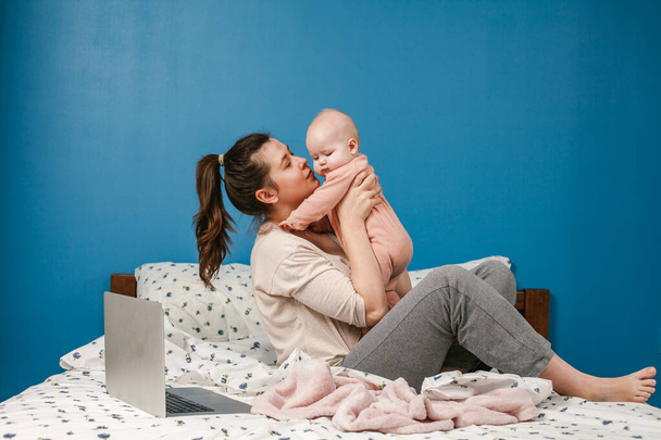 Nuori äiti, jolla on pieni vauva rentoutumassa ja leikkimässä sängyssä. Nuori äiti kotona toimistossa kannettavan tietokoneen ja hänen vastasyntyneen vauvan kanssa. Lapset ja vempaimet. Mukavuutta kotona. Huolellisuutta ja huomiota. Töitä kotoa. Naisten liiketoiminta
. - Valokuva, kuva