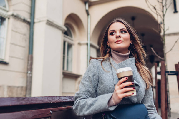 Young stylish woman drinking coffee sitting on bench on city street waiting for someone. Girl relaxing outdoors. Spring female fashion - Foto, Imagen