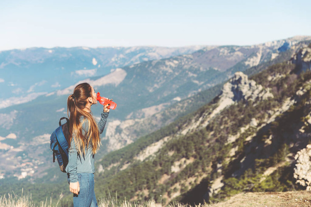 Jong mooi meisje reist alleen in de bergen in het voorjaar of de herfst, kijkt in de verte en geniet van de natuur, rotsen en groene bossen, uitzicht op het landschap. een rugzak achter en sportkleding - Foto, afbeelding