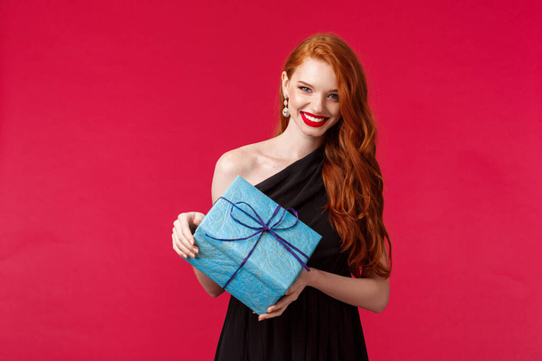 Celebration, holidays and women concept. Portrait of gorgeous young caucasian woman with ginger hair, wear red lipstick black evening dress, look camera happy smiling, holding birthday gift - Fotografie, Obrázek