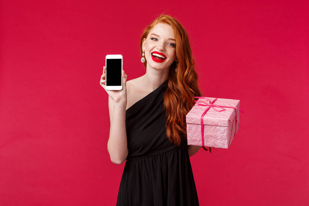 Portrait of gorgeous redhead woman in stylish black dress, laughing and smiling, red lipstick, showing mobile phone application, online show in smartphone display, hold gift box, red background - Photo, Image