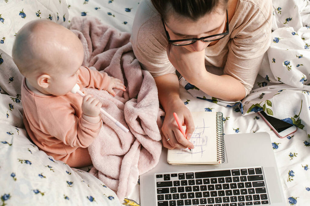 Young mother with her little baby relaxing and playing in the bed. Young mother in home office with laptop and her newborn baby. Children and gadgets. Home comfort. Care and attention. Work from home. Female business. - Photo, Image