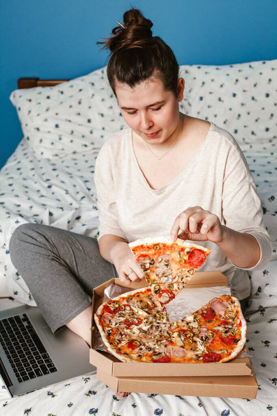 Μια γυναίκα δουλεύει με ένα λάπτοπ στο κρεβάτι και τρώει πίτσα. Fast food, πίτσα έννοια. - Φωτογραφία, εικόνα