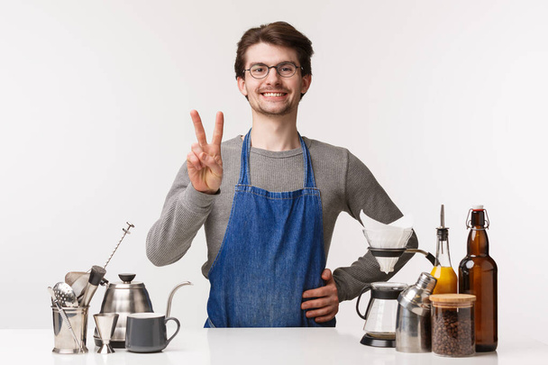 Barista, cafémedewerker en barman concept. Portret van leuke jonge mannelijke werknemer in schort tonen vrede teken en glimlachen, groeten klanten, klaar om te nemen orde, het maken van koffie, thee - Foto, afbeelding