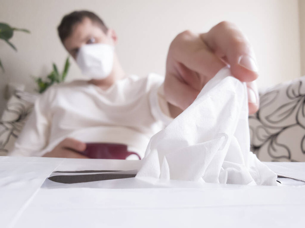 Het uitbreken van de griep of een verkoudheid. Jonge blanke man met een masker op het gezicht reikend naar een wegwerpdoek in de doos. Close-up weefseldoos - Foto, afbeelding
