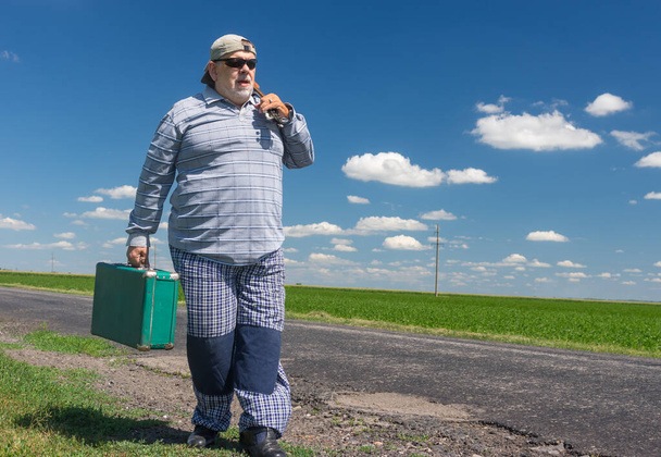 Vousatý starší muž v černých slunečních brýlích se zeleným starobylým kufříkem v pravé ruce a mandolínou na levém rameni chůzi po venkovské silnici  - Fotografie, Obrázek