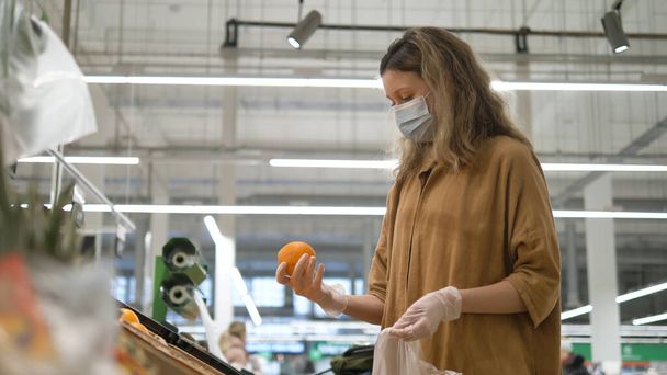 Жінка в медичній масці і рукавички вибирає апельсини в продуктовому супермаркеті. Захист від коронавірусної епідемії, підвищений імунітет зі свіжими фруктами
. - Фото, зображення
