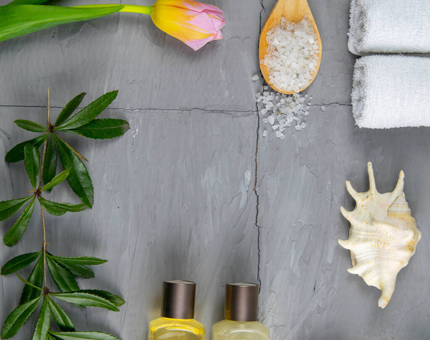 Two towels, body care oil, tulips, green branch and sea salt stand on a concrete background. Spa treatments concept, healthy body. - Photo, Image