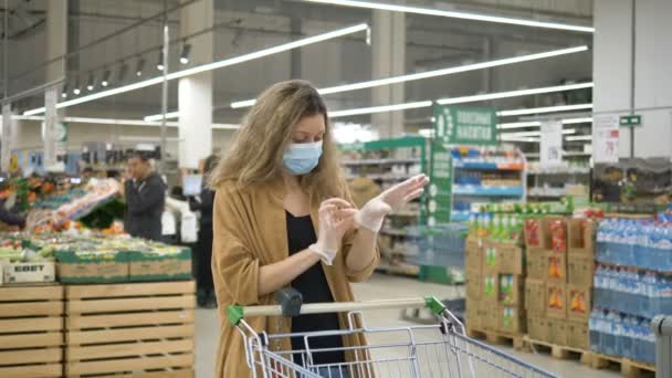 医療用マスクの若い女性は、スーパーマーケットに食料品店のカートで立って、慎重に彼女の手にゴム手袋を調整します。コロナウイルスからの保護. - 映像、動画